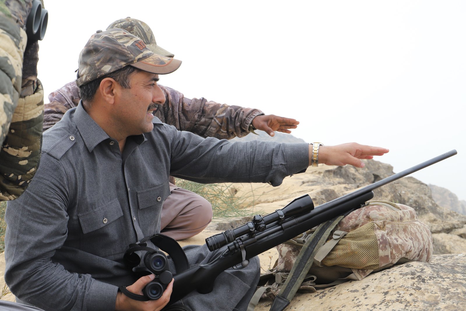 Glassing the ground - a hunting rifle in hand, binoculars ready, western mountains beyond.